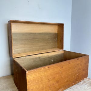 Puede incluir: Una caja de almacenamiento rectangular de madera con una tapa con bisagras en posición abierta. La caja presenta un acabado de grano de madera marrón claro natural con signos visibles de desgaste en las superficies exteriores y un tono más claro.