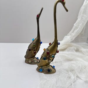 May include: Two brass swan figurines with colorful jewel accents. The swans have long necks and are adorned with red, blue, green, and yellow gems. The bases feature additional jewel details, creating a decorative and elegant aesthetic.