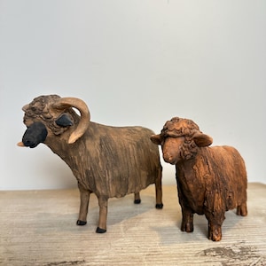 May include: Two hand-crafted clay figurines of sheep standing side by side. The larger sheep on the left features curved horns, a dark brown face, and a light brown textured coat. The smaller sheep on the right has a dark brown textured coat.