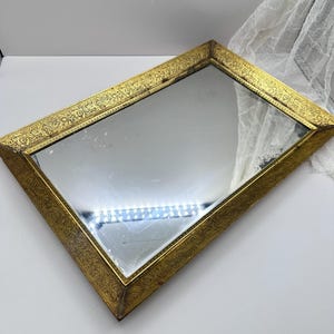Op de afbeelding: Rechthoekige spiegel met een goudkleurige, sierlijke lijst. De lijst heeft een gedetailleerd, herhalend patroon. Het oppervlak van de spiegel reflecteert licht en het ontwerp suggereert een vintage of antieke esthetiek.