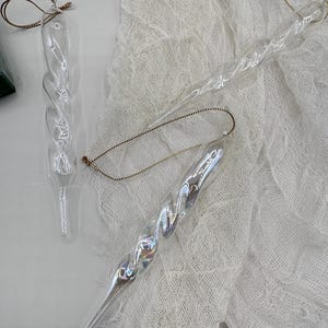 May include: Three clear glass icicle decorations with a twisted design and gold string hangers. The ornaments are displayed on a white, textured fabric background. The icicles have a slight iridescent sheen.