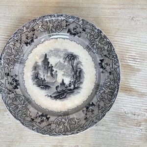 May include: A round ceramic plate featuring a black and white transferware scene of a castle, trees, and figures. The center has a cream-colored background with a crackled glaze, while the wide border displays an intricate floral scroll pattern.