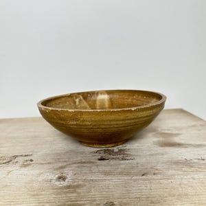 May include: A shallow, circular ceramic bowl with a mottled, earthy brown glaze. The surface features subtle horizontal ridges and variations in color tone, ranging from light tan to deep ochre, with a smooth, slightly reflective finish.