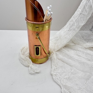May include: A copper and brass match holder with a curved top and a handle. The cylindrical container holds several matches with white tips. The holder has brass bands around the top and bottom.