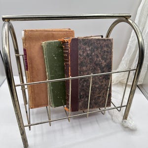 May include: A vintage metal magazine rack holding three antique books. The books have aged covers in shades of brown and green. The rack is made of a silver-toned metal with a rectangular shape and curved sides.