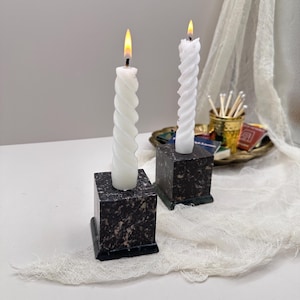Op de afbeelding: Twee brandende witte spiraalkerstjes staan op donkere, gespikkelde vierkante marmeren kandelaars. De kaarsvlammen zijn geel. De houders staan op een wit oppervlak met een decoratieve doek.