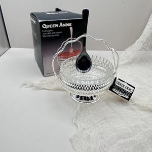 May include: A Queen Anne jam dish with a silver-colored handle and base, and a clear glass bowl. A black spoon rests inside the bowl. The product is in its original box, which is black with white text. A white cloth is draped in the background.
