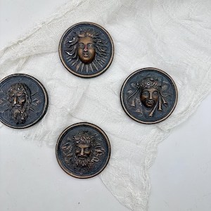 May include: Four antique bronze-colored medallions with detailed relief portraits. Each circular medallion features a different classical face, including a sun god, a bearded man, and a woman with a laurel wreath. The medallions are arranged on a white, textured cloth.