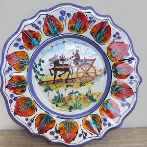 Peut inclure: Une assiette décorative en céramique avec un bord festonné. L'assiette présente une scène centrale d'une charrette tirée par des chevaux et une bordure de formes stylisées en forme de feuilles rouges et oranges. Le bord est de couleur bleu foncé.