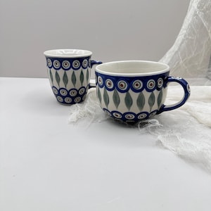May include: Two ceramic mugs with a blue and white pattern. The mugs have a white interior and a blue rim. The pattern includes green leaf shapes and red and blue circles. The mugs have a handle.