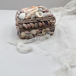 Puede incluir: Una caja rectangular cubierta de varias conchas marinas, con un borde similar a una cuerda. La caja está adornada con una variedad de conchas en tonos blancos, marrones y beige. Es un objeto decorativo.