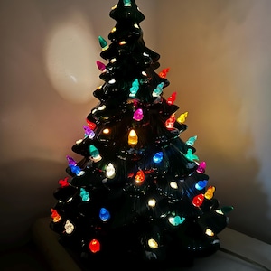 Könnte beinhalten: Ein dunkelgrüner Weihnachtsbaum aus Keramik mit glänzender Oberfläche, der mit zahlreichen kleinen, bunten Kunststoff-Lichtern in Rot, Blau, Gelb und Türkis verziert ist, die von innen heraus beleuchtet werden.