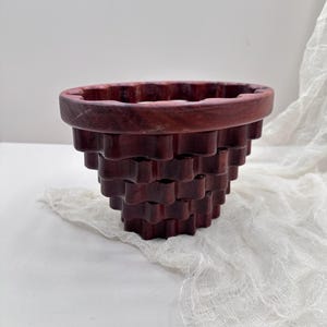 Può includere: Una ciotola decorativa in legno rosso scuro con un design unico a strati. La ciotola ha un bordo ampio e arrotondato e un motivo geometrico strutturato sui lati. La ciotola è probabilmente fatta a mano e adatta all'arredamento della casa.