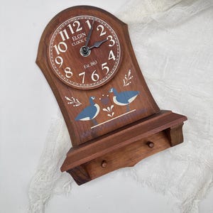 Puede incluir: Reloj de pared de madera con acabado marrón. La esfera del reloj tiene números y manecillas blancas, con el texto "ELGIN CLOCK CO." y "Est. 1863". El reloj presenta una repisa con dos clavijas y un diseño pintado de pájaros azules.