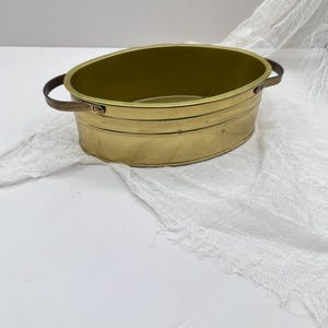 Puede incluir: Un pequeño recipiente ovalado de latón con dos asas marrones oscuras. El recipiente tiene un interior y exterior dorados, con líneas horizontales alrededor de los lados. El fondo es una superficie blanca con una tela blanca texturizada.