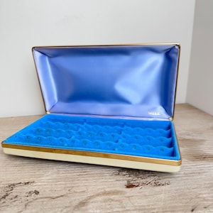 Puede incluir: Joyero abierto con exterior crema y ribete dorado. El interior presenta un forro de satén azul vibrante y una bandeja de terciopelo azul con ranuras para anillos. La marca "MELE" es visible dentro de la tapa.