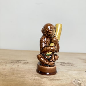 May include: A small, glossy brown ceramic figurine of a monkey sitting on a circular base. The monkey is depicted in a seated position, holding a smooth, pale yellow cylindrical object against its shoulder with both hands.