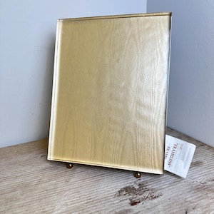 May include: A rectangular frameless glass display frame with a gold-toned finish and a subtle wood grain texture. The frame stands on two small gold-colored spherical feet and includes a white tag labeled THE "FRAMELESS" FRAME.