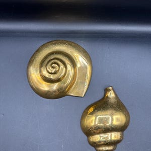 Puede incluir: Dos esculturas de conchas marinas de latón. Una tiene forma de espiral y la otra de concha. Las esculturas tienen un acabado dorado brillante. Estas piezas decorativas son adecuadas para una decoración de temática costera o náutica.