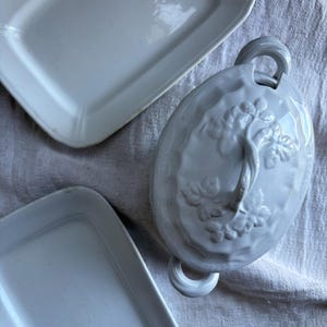 May include: A set of three white ceramic dishes. The largest dish has a raised floral design and two handles. The two smaller dishes are rectangular.