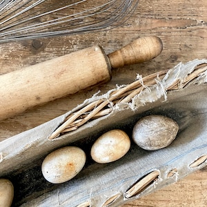 May include: A rustic kitchen scene featuring a vintage wire whisk, a wooden rolling pin, and several speckled eggs nestled in a distressed linen trough. The items rest on a weathered wooden surface, evoking a sense of old-world charm.