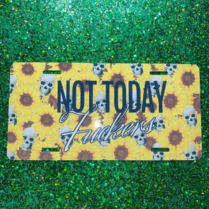 May include: A rectangular license plate with a yellow sunflower background and white skull accents. The plate features the text "NOT TODAY Fuckers" in a bold, stylized font. The plate has four mounting holes.