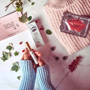 May include: A white and gold tube of eyelash serum with the text "Natura Lashes" and "Filler Enhancing Serum" on the label. The tube is on a white marble surface with a pink sweater, a silver lip mask, and green leaves.