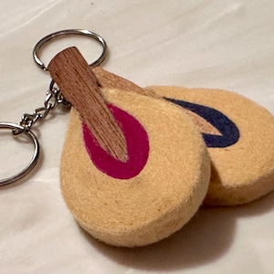 May include: A piano hammer keychain featuring two beige felt hammer heads with a pink and blue accent. The hammers have a wooden handle and are attached to a silver keyring. Musical instrument themed.