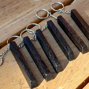 May include: A collection of seven dark brown wooden keychains with silver-colored key rings and chains. The keychains are rectangular with beveled edges, arranged on a light brown wooden surface.