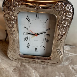 May include: A silver-toned clock with a white face and Roman numeral hour markers. The clock has a decorative frame with floral designs.