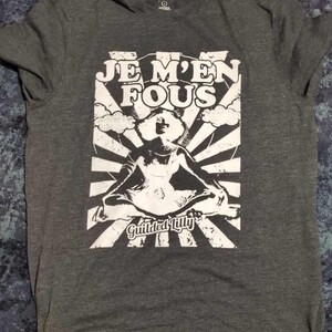 Puede incluir: Una camiseta gris oscuro con un estampado gráfico blanco de una mujer sentada en posición de loto con el texto "Je M'en Fous" y "Gilded Lilly" debajo.