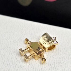 Puede incluir: Encanto de robot dorado con un lazo rosa. El robot tiene un corazón en el pecho y una cara sonriente.