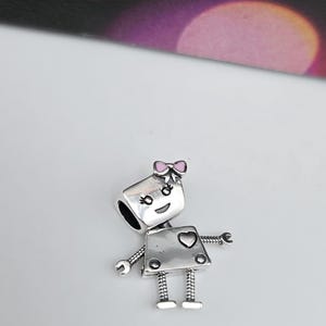 Puede incluir: Un charm de plata con forma de robot con un lazo rosa, un detalle en forma de corazón y pequeños brazos y piernas. El charm probablemente está hecho de plata de ley y está diseñado para ser añadido a una pulsera o collar. Mide aproximadamente 2 cm de altura.