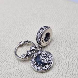 May include: A silver charm featuring a blue and white moon and stars design. The charm has a small ring at the top and is adorned with clear cubic zirconia stones. The charm is marked with the text "S 925 ALE".