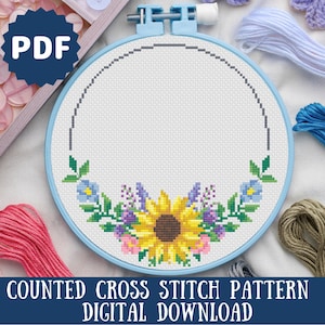 Puede incluir: Un aro de bordado azul con un paño Aida blanco en su interior. El paño tiene un patrón de punto de cruz de un girasol con flores azules y moradas alrededor. El texto "COUNTED CROSS STITCH PATTERN DIGITAL DOWNLOAD" está en la parte inferior de la imagen.