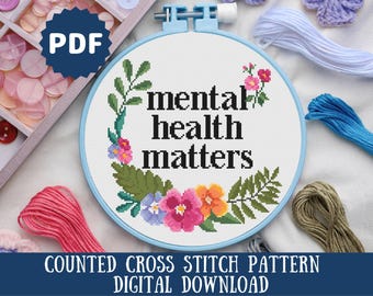 以下が含まれることがあります： 花輪と「mental health matters」というテキストが描かれたカウントクロスステッチパターン。デジタルダウンロードとして入手できます。