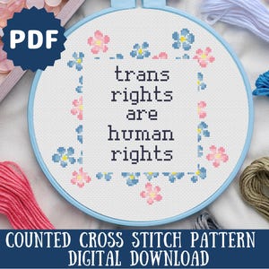 May include: A cross-stitch embroidery hoop with the text "trans rights are human rights." The design is surrounded by pink and blue floral accents. The words "COUNTED CROSS STITCH PATTERN DIGITAL DOWNLOAD" are at the bottom.