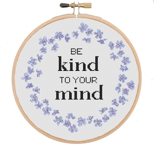 Peut inclure: Un cercle de broderie blanc avec un motif floral bleu autour du bord. Le texte "Be kind to your mind" est centré en noir.
