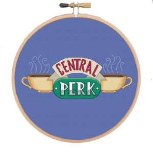 May include: A blue and white embroidery hoop with a cross-stitch design of the "Central Perk" logo from the TV show "Friends". The logo features two coffee cups with steam rising from them and the words "Central Perk" in a green and red font.