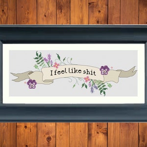 May include: A cross-stitch embroidery with a banner that reads "I feel like shit" surrounded by purple, pink, and green flowers.