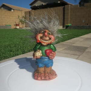 Puede incluir: Una figurita de troll de pelo gris con una camisa verde, pantalones cortos azules y un coraz&oacute;n rojo en una mano y una bola dorada en la otra. El troll tiene una gran sonrisa y est&aacute; de pie sobre una superficie blanca.