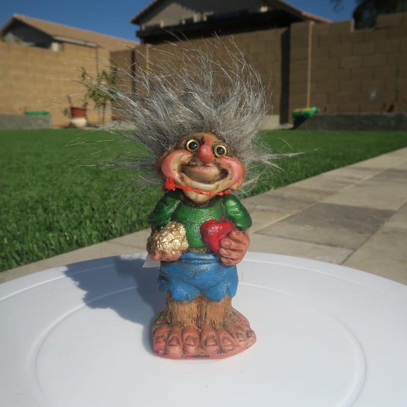 Puede incluir: Una figurita de troll de pelo gris con una camisa verde, pantalones cortos azules y un coraz&oacute;n rojo en una mano y una bola dorada en la otra. El troll tiene una gran sonrisa y est&aacute; de pie sobre una superficie blanca.