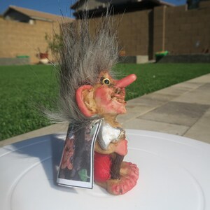 Pu&ograve; includere: Una piccola statuetta di troll con i capelli grigi, che indossa un mantello rosso e nero, tiene un'ascia e un bastone. Il troll ha un naso grande e un sorriso malizioso.