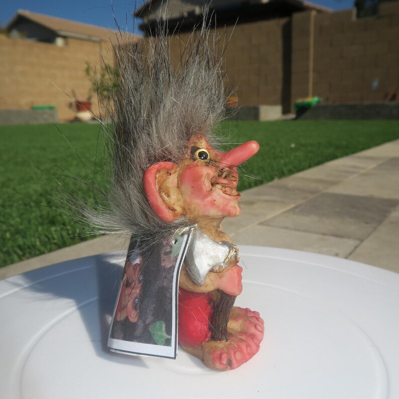 Pu&ograve; includere: Una piccola statuetta di troll con i capelli grigi, che indossa un mantello rosso e nero, tiene un'ascia e un bastone. Il troll ha un naso grande e un sorriso malizioso.