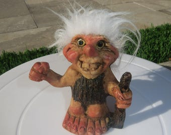 Puede incluir: Una figura de troll vintage con una expresión sonriente, cabello blanco y nariz roja. El troll sostiene un objeto marrón oscuro en su mano derecha y tiene un atuendo negro y marrón. La figura está sobre una superficie blanca.