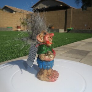 Puede incluir: Una peque&ntilde;a figura de troll de pelo gris que lleva una camisa verde, pantalones cortos azules y sostiene un coraz&oacute;n rojo. El troll tiene una nariz grande y mira hacia la derecha. La figura est&aacute; sentada sobre una superficie blanca.