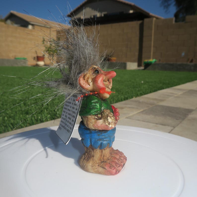 Puede incluir: Una peque&ntilde;a figura de troll de pelo gris que lleva una camisa verde, pantalones cortos azules y sostiene un coraz&oacute;n rojo. El troll tiene una nariz grande y mira hacia la derecha. La figura est&aacute; sentada sobre una superficie blanca.