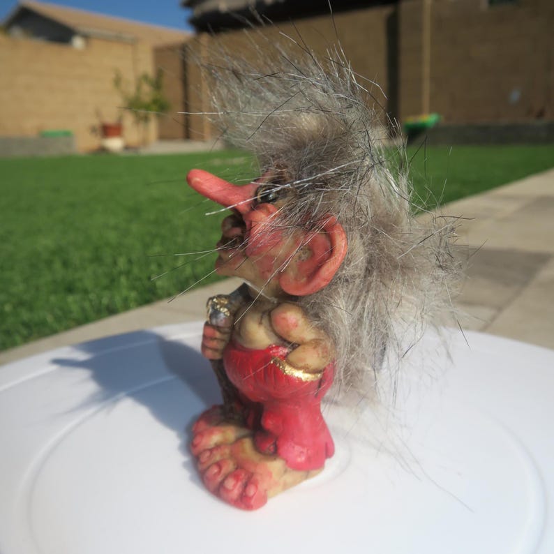 Pu&ograve; includere: Una piccola statuina di troll con i capelli grigi, con un naso lungo, che indossa un perizoma rosso e oro e tiene un bastone.