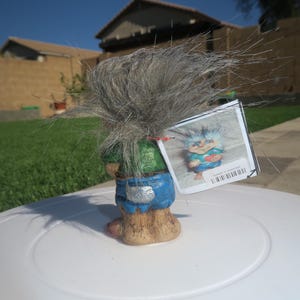 Puede incluir: Una figurita de troll de pelo gris que lleva un atuendo verde y azul. El troll sostiene un coraz&oacute;n rojo en su mano. La figurita est&aacute; de pie sobre una superficie blanca. La figurita tiene una etiqueta adjunta que dice "Edici&oacute;n limitada".