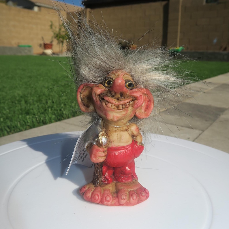 Pu&ograve; includere: Una piccola statuina di troll con i capelli grigi, che indossa un perizoma rosso e un ampio sorriso. Il troll tiene un bastone di legno con un anello d'oro in cima.